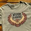 Front of Livin' Light's "Wisdom Comes from Above" olive green, short-sleeved T-shirt with tree background. Owl with wings spread and piercing eyes says Wisdom Comes From Above which illustrates Bible verse Proverbs 2:6.