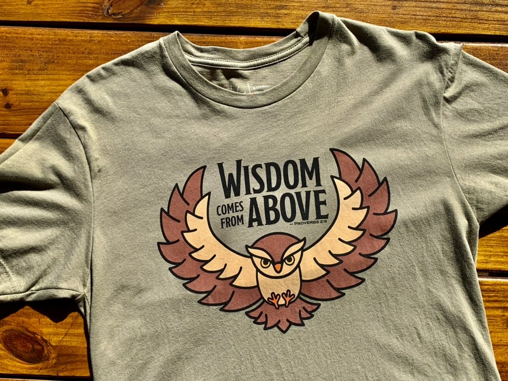 Front of Livin' Light's "Wisdom Comes from Above" olive green, short-sleeved T-shirt with tree background. Owl with wings spread and piercing eyes says Wisdom Comes From Above which illustrates Bible verse Proverbs 2:6.