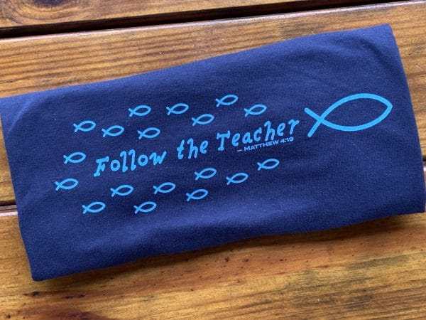 Close up of front of Livin' Light's "Follow the Teacher" T-shirt on wood background. On a short-sleeved, navy shirt, a school of light blue fish are following the big fish, Jesus with the word's "Follow the Teacher" in the middle of the school. Matthew 4:19.