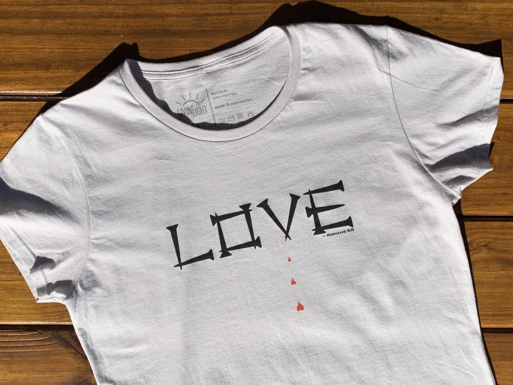 LOVE front, wood background: This Livin' Light shirt spells out the word Love in nails. From one nail, blood drips forming into hearts. This bold design on a gray, short-sleeved shirt illustrates Romans 5:8
