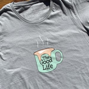 Front of Livin' Light's The Good Life coffee mug shirt with overflowing froth heart on wood background. XL light green mug pops on dark gray shirt. Psalm 23:5-6