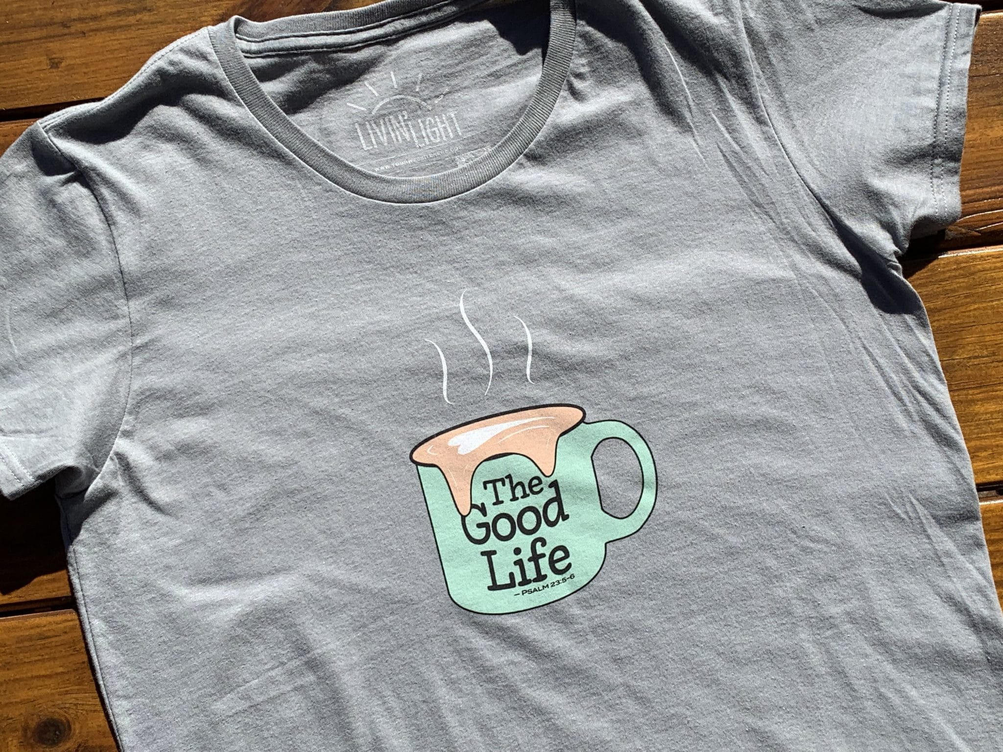 Front of Livin' Light's The Good Life coffee mug shirt with overflowing froth heart on wood background. XL light green mug pops on dark gray shirt. Psalm 23:5-6