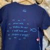 Back of Livin' Light's "Follow the Teacher" T-shirt against stone wall. On a short-sleeved, navy shirt, a school of light blue fish are following the big fish, with Jesus words in Matthew 4:19, "Follow Me and I will make you fishers of people."