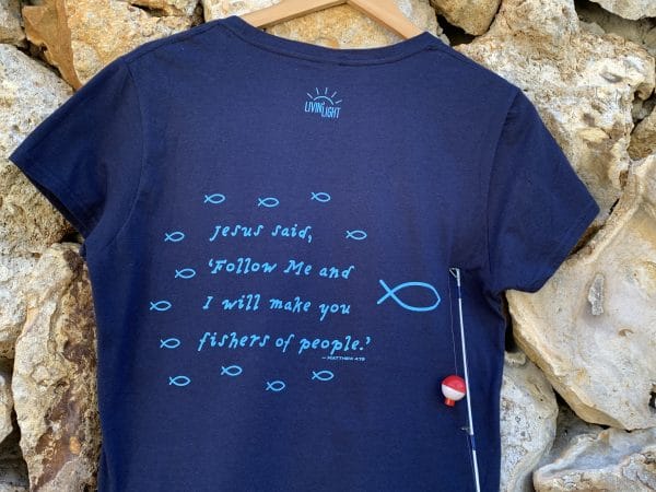 Back of Livin' Light's "Follow the Teacher" T-shirt against stone wall. On a short-sleeved, navy shirt, a school of light blue fish are following the big fish, with Jesus words in Matthew 4:19, "Follow Me and I will make you fishers of people."