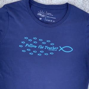 Front of Livin' Light's "Follow the Teacher" T-shirt on sidewalk concrete background. On a short-sleeved, navy shirt, a school of light blue fish are following the big fish, Jesus with the word's "Follow the Teacher" in the middle of the school. Matthew 4:19.