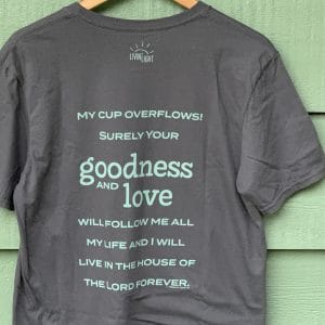 Back of dark gray, short-sleeved, The Good Life shirt on green wood wall. Psalm 23:5-6 says: My cup overflows! Surely your goodness and love will follow me all my life and I will live in the house o the Lord forever.