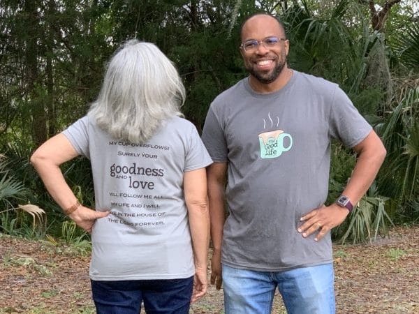 Female and male models showing light gray and medium gray versions of Livin' Light's The Good Life T-shirt. Models also showing the front and back. Overflowing mug on front at a park. Psalm 23:5-6 on back.