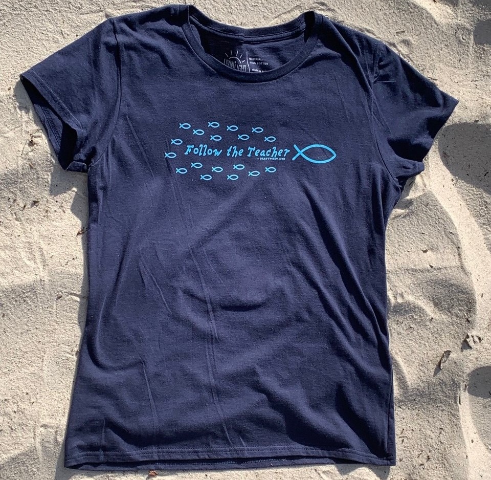 Front of Livin' Light's "Follow the Teacher" T-shirt on sand background. On a short-sleeved, navy shirt, a school of light blue fish are following the big fish, Jesus with the word's "Follow the Teacher" in the middle of the school. Matthew 4:19.